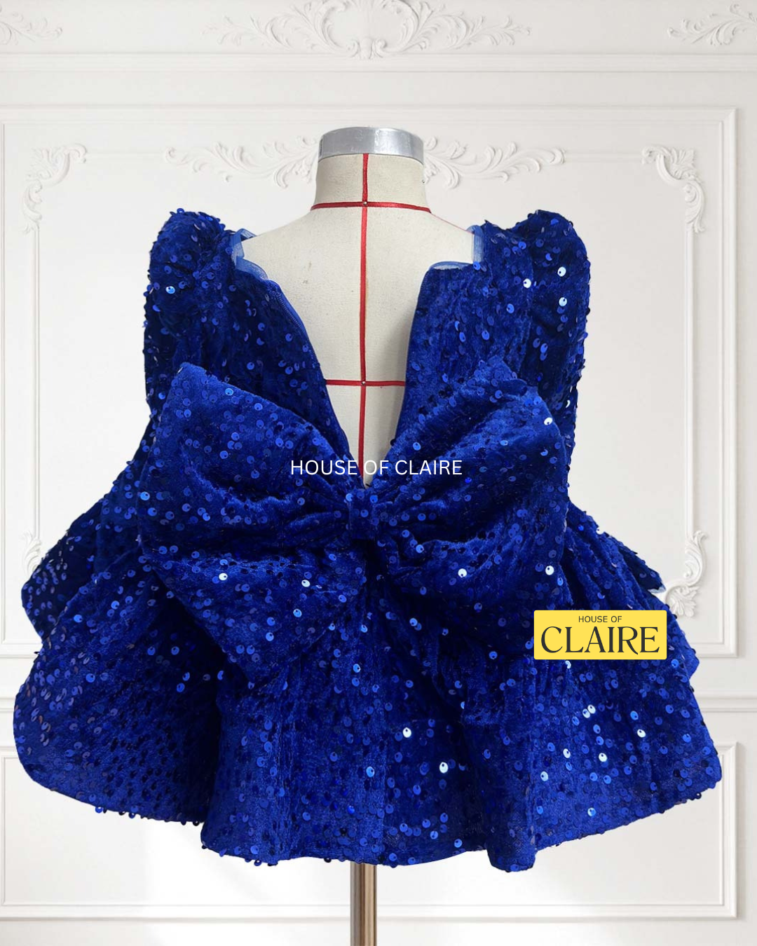 A royal blue sequined children's dress by house of claire displayed on a mannequin against a white decorative wall. The dress has a square neckline, puffed sleeves, and a tiered ruffled skirt. www.houseofclaire.com