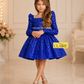 A young girl stands in a bright indoor room wearing a royal blue sequined dress with long puff sleeves, a square neckline, and a flared skirt by house of claire kids dresses bangalore.She is wearing silver shoes and a headband. www.houseofclaire.com