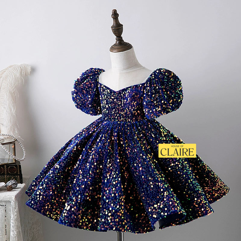 A navy blue sequined dress with puff sleeves and a sweetheart neckline displayed on a mannequin. The dress is covered in iridescent sequins that reflect multiple colors.Navy blue sequin baby girl party dress with puff sleeves, sheer neckline and full skirt for birthday parties - house of claire kids party wear dress bangalore online india