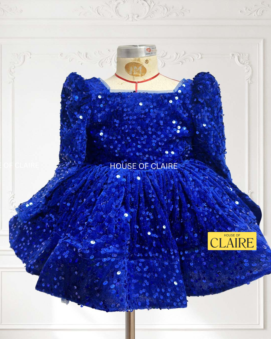 A royal blue sequined children's dress by house of claire displayed on a mannequin against a white decorative wall. The dress has a square neckline, puffed sleeves, and a tiered ruffled skirt. www.houseofclaire.com