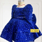 A royal blue sequined children's dress by house of claire displayed on a mannequin against a white decorative wall. The dress has a square neckline, puffed sleeves, and a tiered ruffled skirt. www.houseofclaire.com