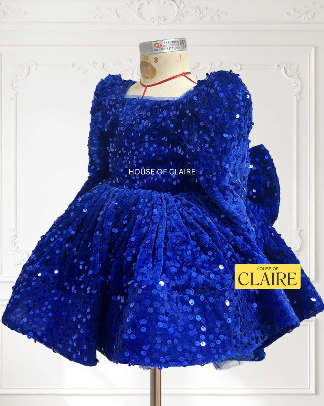 A royal blue sequined children's dress by house of claire displayed on a mannequin against a white decorative wall. The dress has a square neckline, puffed sleeves, and a tiered ruffled skirt. www.houseofclaire.com